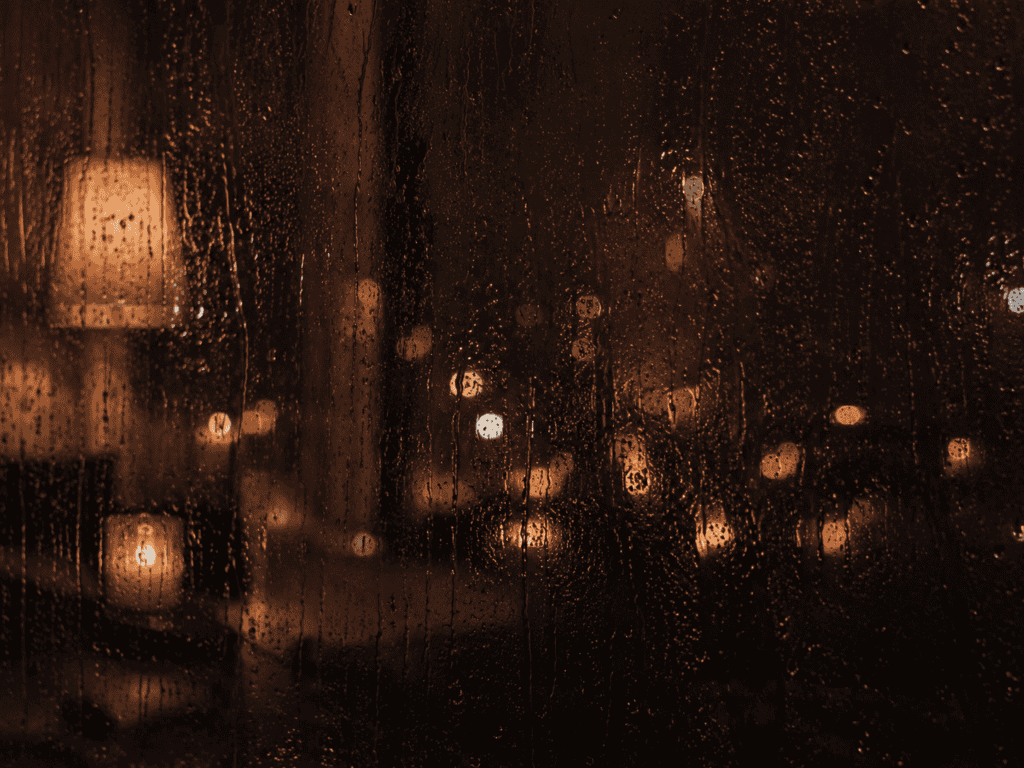 Rain-covered window at night with blurred warm lights and reflections glowing through the glass.